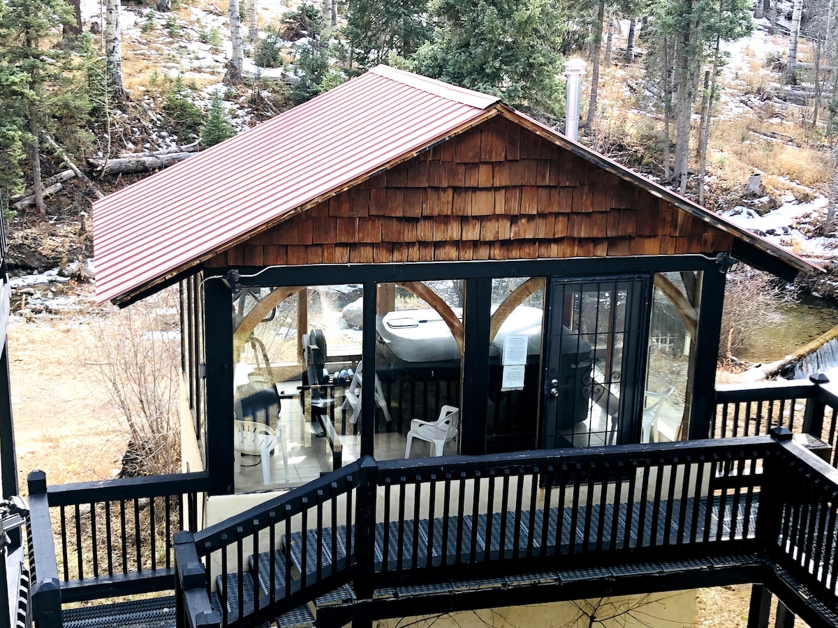 A charming outdoor building is situated near the forest, featuring large glass windows that provide views of the surrounding trees. The structure includes a hot tub, and a small deck with chairs is visible, allowing for outdoor relaxation in a serene environment.