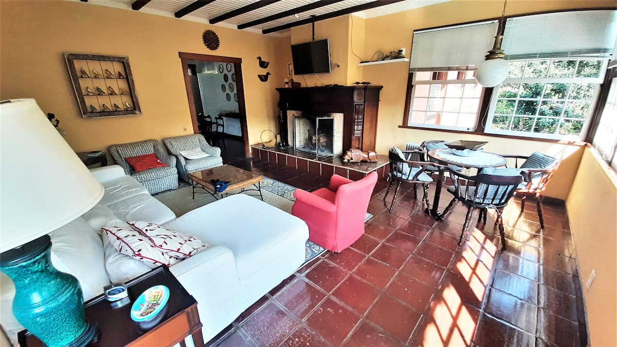 A spacious living area is captured, featuring a cozy seating arrangement with a white sofa and armchairs in soft fabrics. A wooden fireplace serves as a focal point, while a dining table sits nearby, complemented by large windows allowing natural light to illuminate the room.