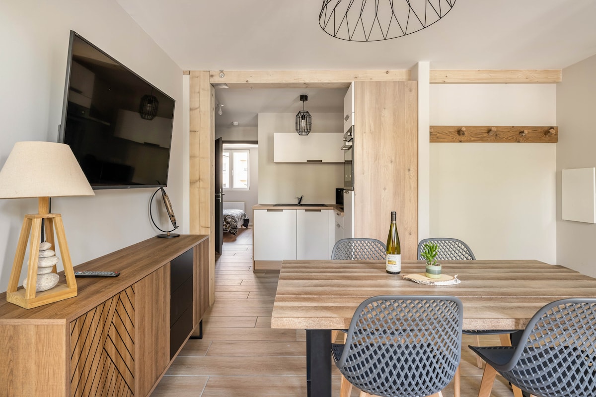 A dining area showcases a wooden table surrounded by four modern chairs. A minimalist lamp and a bottle of wine add to the setting. A large television is mounted on the wall, while the open kitchen and living space are visible in the background.