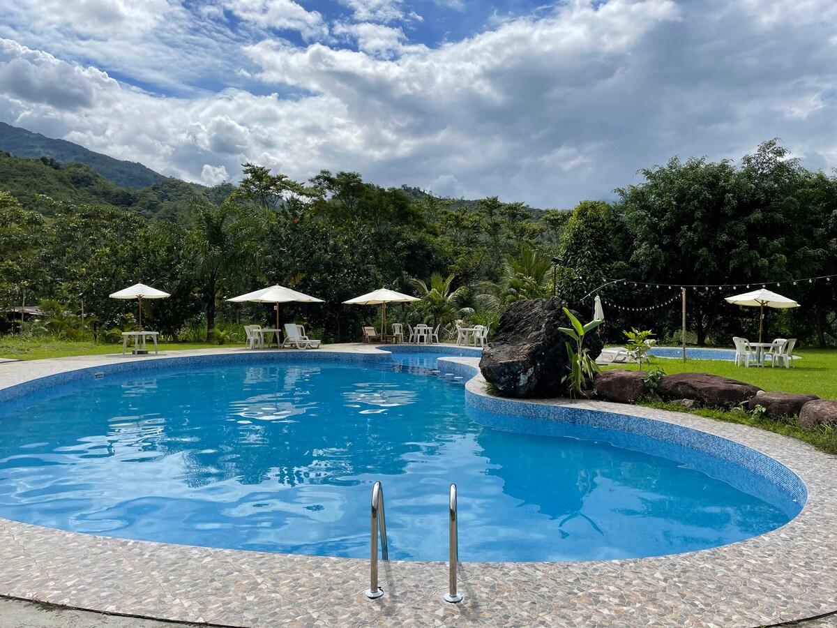 A serene pool area is surrounded by lush greenery, featuring an inviting blue water surface and gentle stone detailing. Umbrellas and white chairs are positioned around the pool, providing ample seating. A natural rock formation adds interest to the landscape, with mountains visible in the distance.