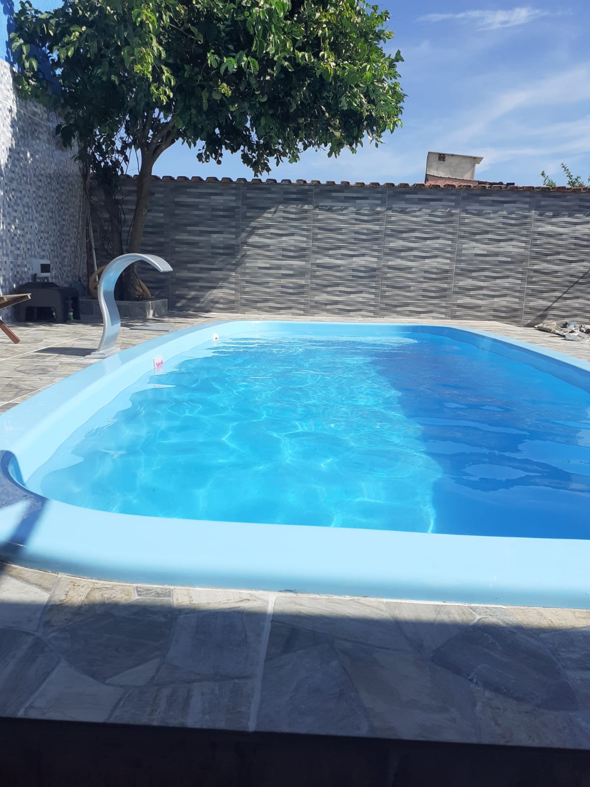 A serene outdoor pool is surrounded by gray stone tiles and a low wall. Sunlight reflects off the clear water, creating a soothing ambiance. A tree provides partial shade, contributing to the tranquil environment.