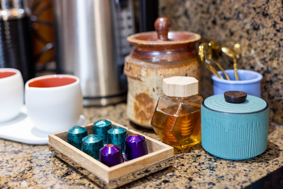 A well-arranged kitchen counter features a variety of visually appealing items. Two white cups with red interiors sit alongside a wooden tray holding six coffee capsules. A honey jar with a wooden lid and a turquoise container are also present, adding a touch of color to the granite surface.