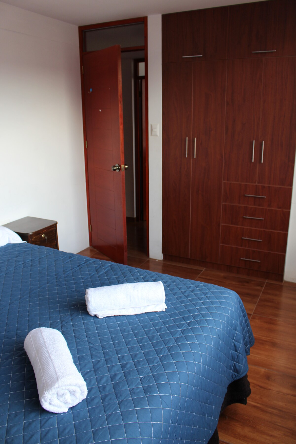 A spacious bedroom features a comfortable bed covered with a blue quilt and two rolled towels placed on top. A wooden wardrobe with multiple drawers is present against the wall, and a door leads to another room, enhancing the room's functionality.