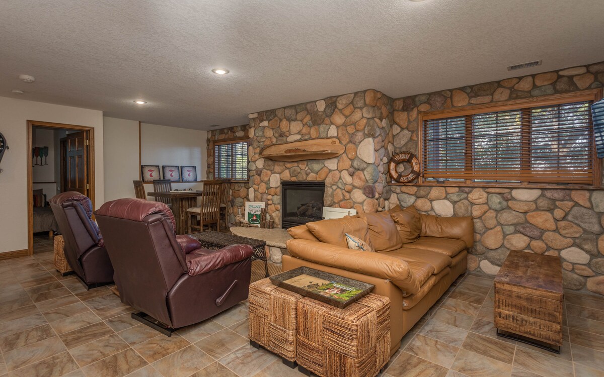 A cozy living area features a stone wall and a fireplace, accompanied by a medium-sized brown leather sofa and two recliner chairs. Natural light enters through the two partially covered windows, and a wooden dining table is visible in the background.
