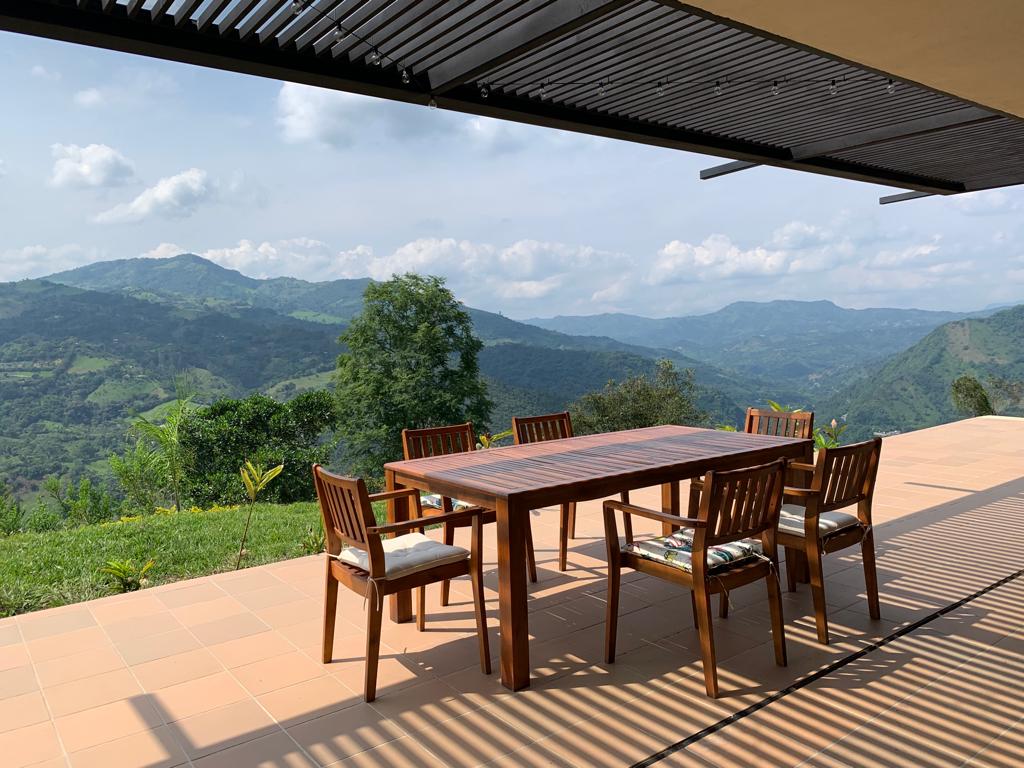 A spacious outdoor terrace features a wooden dining table surrounded by six chairs. The breathtaking view of the mountains and lush landscape is visible in the background, with an expansive sky overhead.
