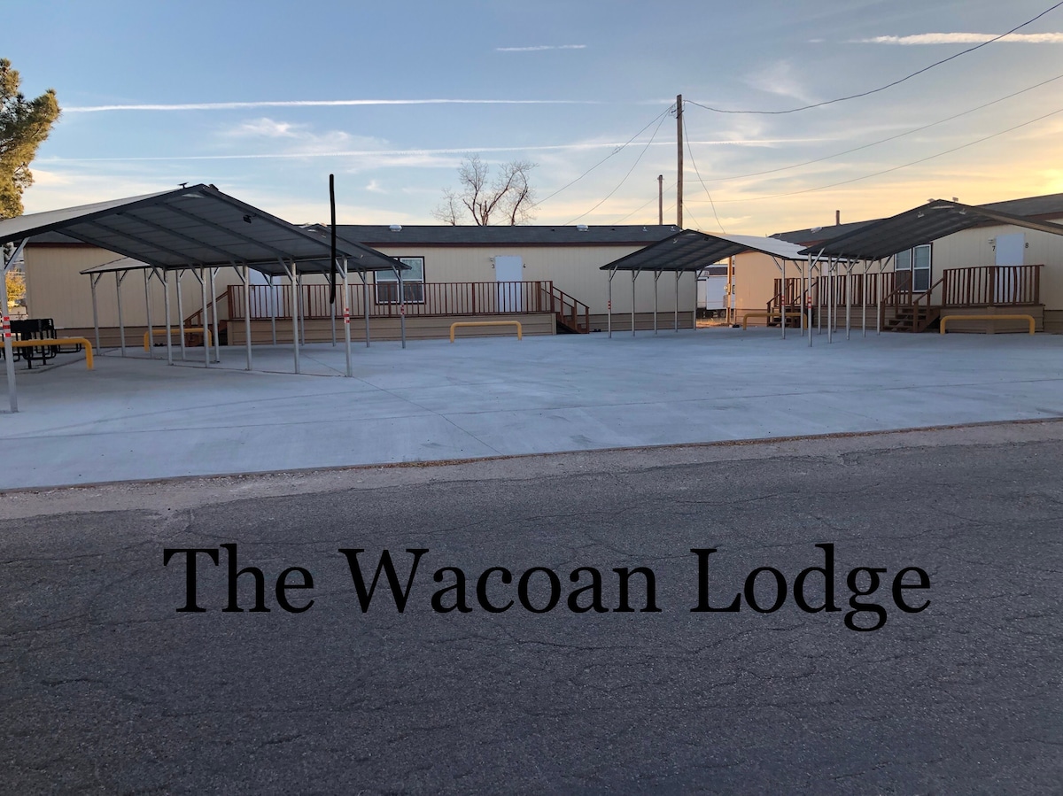 The exterior of the Wacoan Lodge is shown, featuring a spacious parking area with multiple covered parking spots. The property includes two single-story buildings, each with a staircase leading to the entrance. A clear sky is visible in the background.