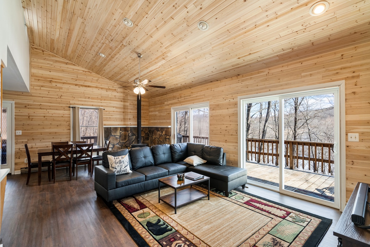 A spacious living area features a dark sectional sofa, complemented by a wooden coffee table and patterned area rug. Large glass doors provide views of the wooded outdoors, while a dining area with a wooden table is visible in the background. Wooden paneling adds warmth to the space.