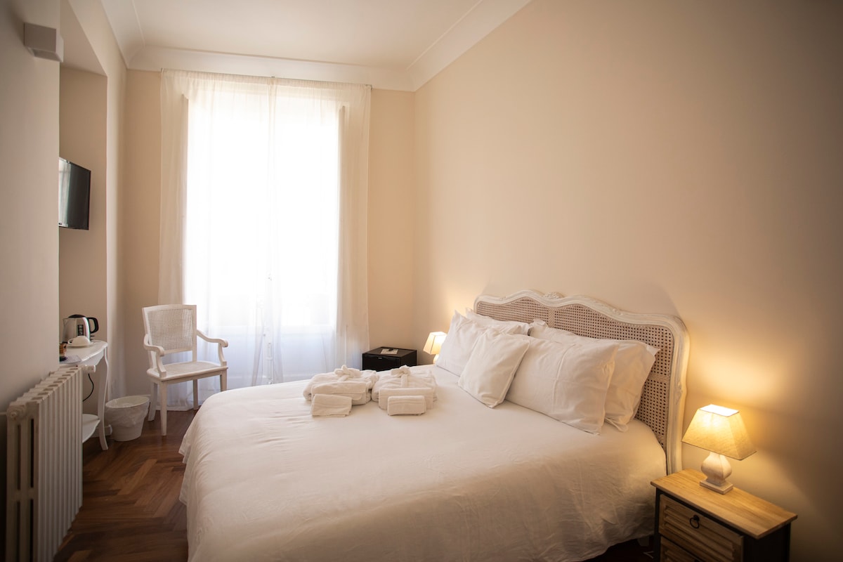 The room features a spacious bed with neatly arranged white linens, complemented by soft lighting from bedside lamps. A minimalist white chair and a small table are positioned near a large window, allowing natural light to fill the space, enhancing its airy feel.