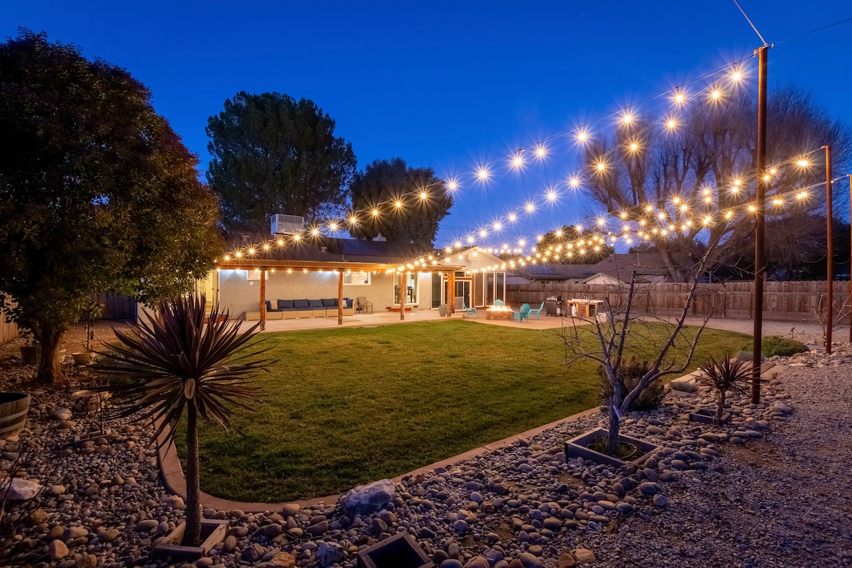 A spacious backyard is illuminated by string lights, creating a warm ambiance at dusk. Green grass covers the lawn, bordered by decorative rock gardens and low shrubs. A covered patio area is visible, providing shaded seating and inviting space for outdoor gatherings.