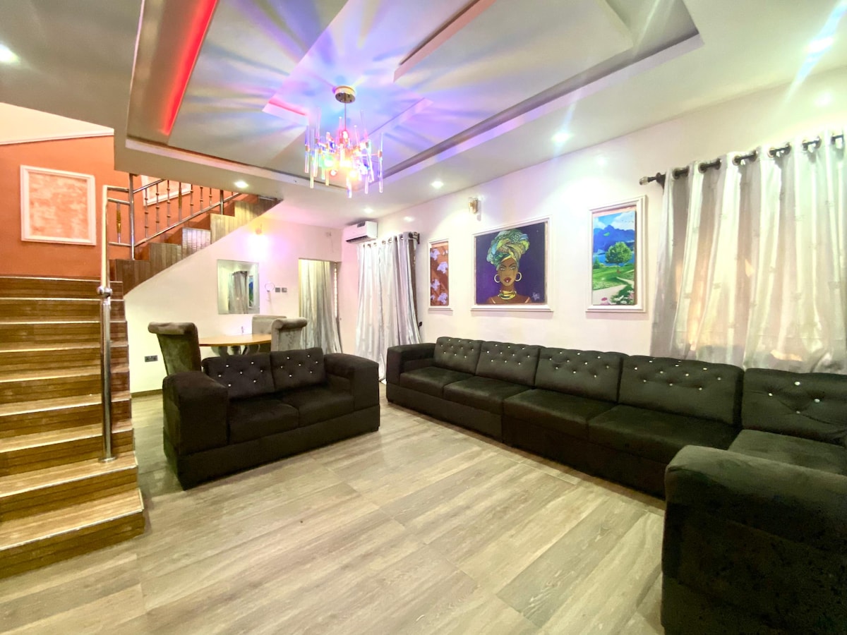 A spacious living area is highlighted by two large, black sofas arranged to promote conversation. The walls are adorned with vibrant artwork. Soft lighting from a modern chandelier creates a relaxed atmosphere. Wooden stairs lead to an upper level, complementing the elegant design.