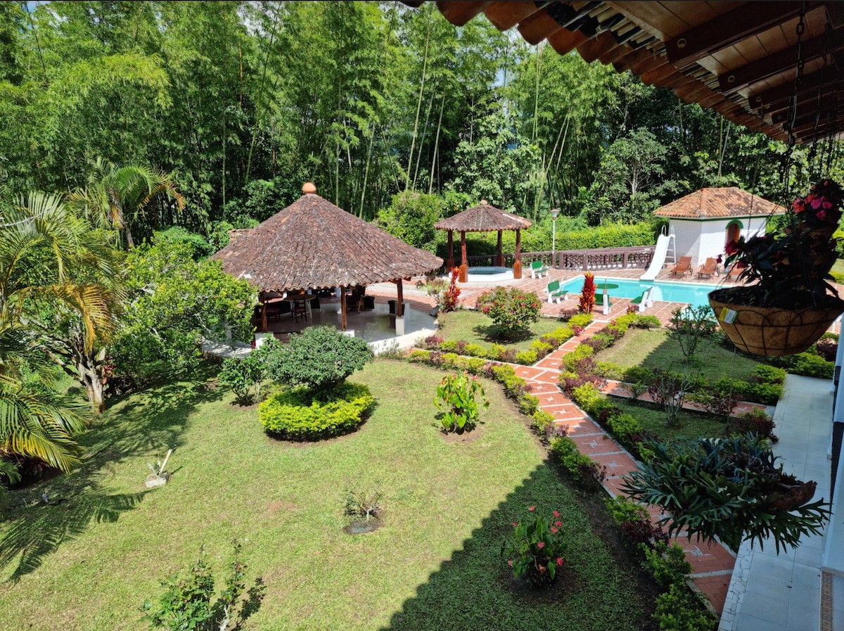 The outdoor area features a lush garden with manicured lawns and vibrant flower beds. A thatched-roof kiosk is visible, along with a turquoise swimming pool surrounded by sun loungers. Pathways meander through the greenery, offering a tranquil setting for relaxation.