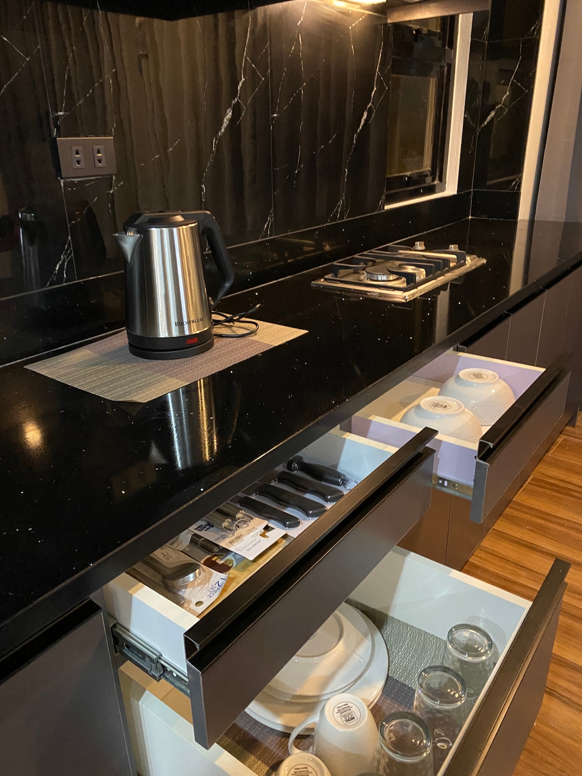 A modern kitchen features sleek black countertops with a kettle and a gas stove visible. Drawers below display a set of dishes and glassware, highlighting functional storage options. Soft lighting accentuates the marble-like backsplash, creating an elegant cooking space.
