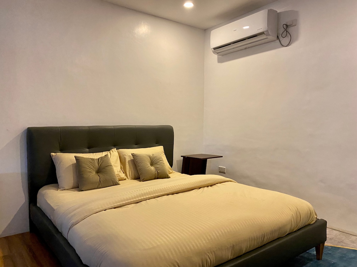 A spacious bed is dressed in neutral-toned linens, accompanied by two decorative pillows in a soft hue. An air conditioning unit is mounted on the wall above, providing climate control, while a small bedside table is positioned to the side.