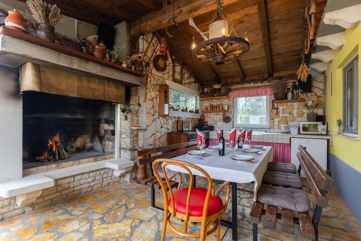 A rustic kitchen area is highlighted by a stone fireplace which adds warmth to the space. A long dining table is set with red napkins, surrounded by wooden benches. Traditional decor elements, including various kitchenware, embellish the walls beneath a wooden ceiling.