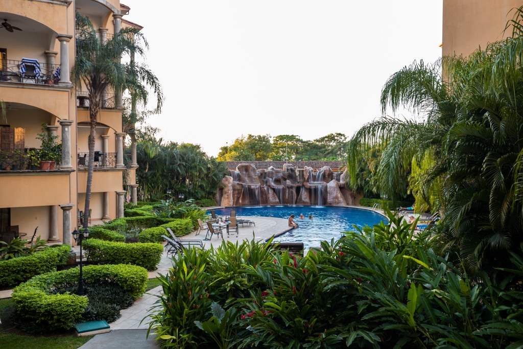A tropical landscape features a large swimming pool surrounded by lush greenery. A stone waterfall cascades into the pool, providing a serene focal point. Lounge chairs are placed around the pool, inviting relaxation, while palm trees frame the scene.