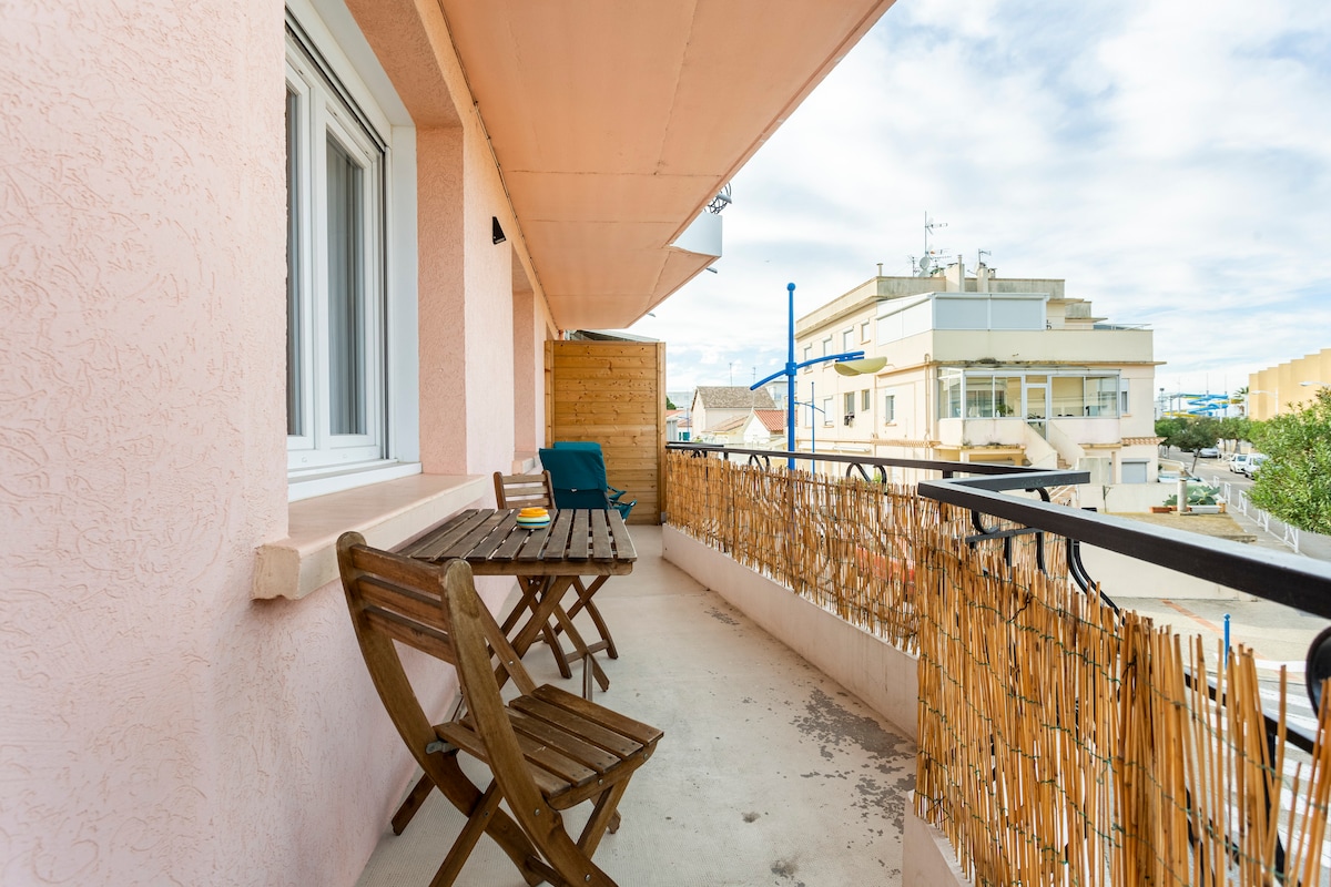 A spacious terrace features a wooden table with two chairs, offering a pleasant outdoor seating area. A bamboo railing provides privacy while allowing for natural light. The surrounding view includes neighboring buildings and an open sky.