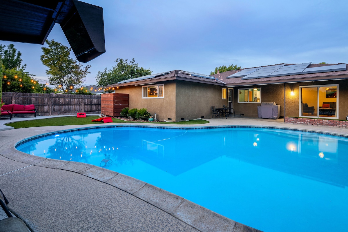 The outdoor space features a well-maintained pool surrounded by a spacious patio area. Comfortable seating options are visible, and landscaping elements provide a relaxing ambiance. The evening sky casts a soft glow, while string lights enhance the welcoming atmosphere.