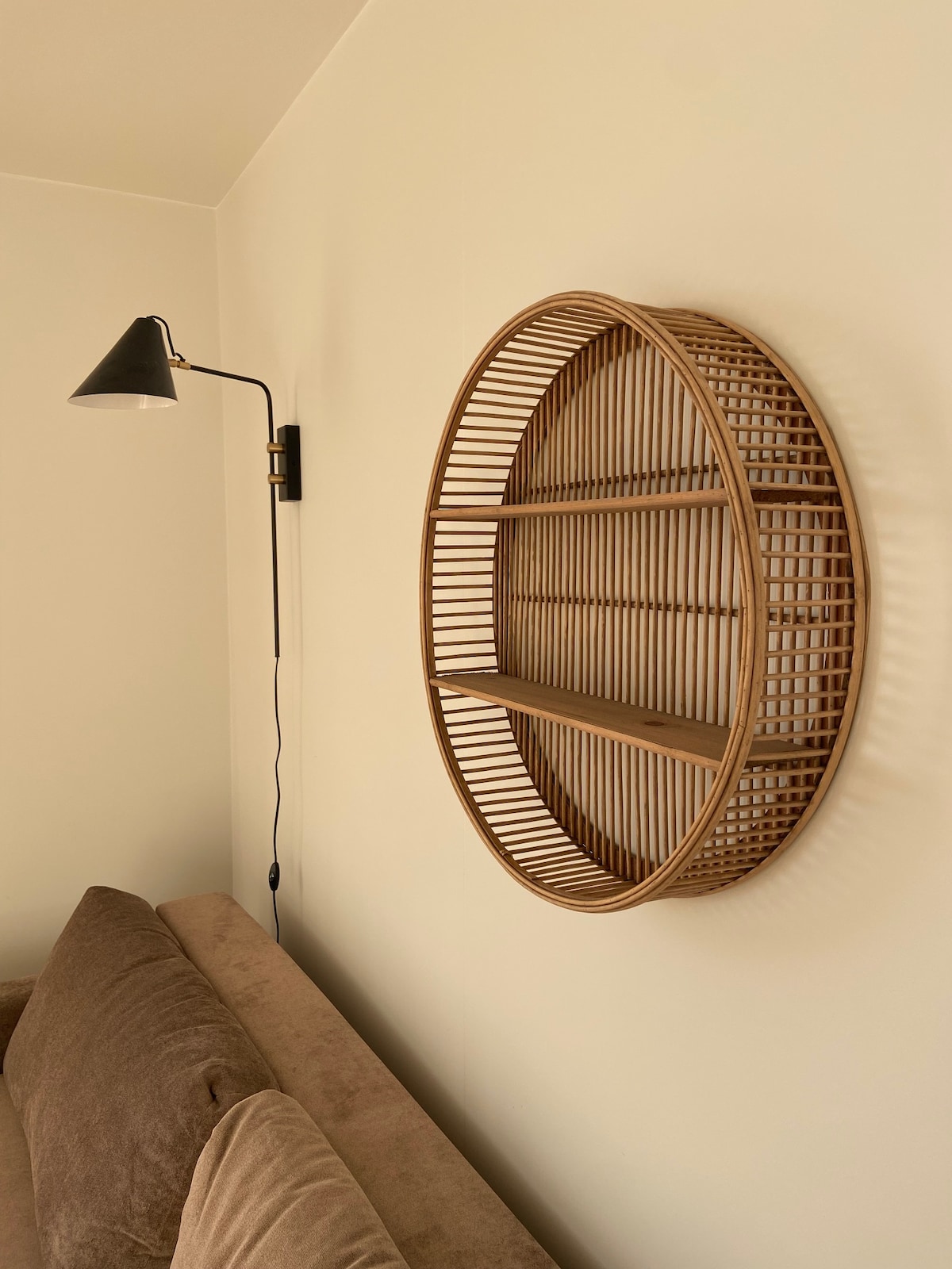 A circular rattan shelf is mounted on the wall, featuring two open levels for decorative items. A wall-mounted lamp is positioned nearby, providing functional lighting. The adjacent sofa is upholstered in a soft brown fabric, complementing the warm tones of the room.