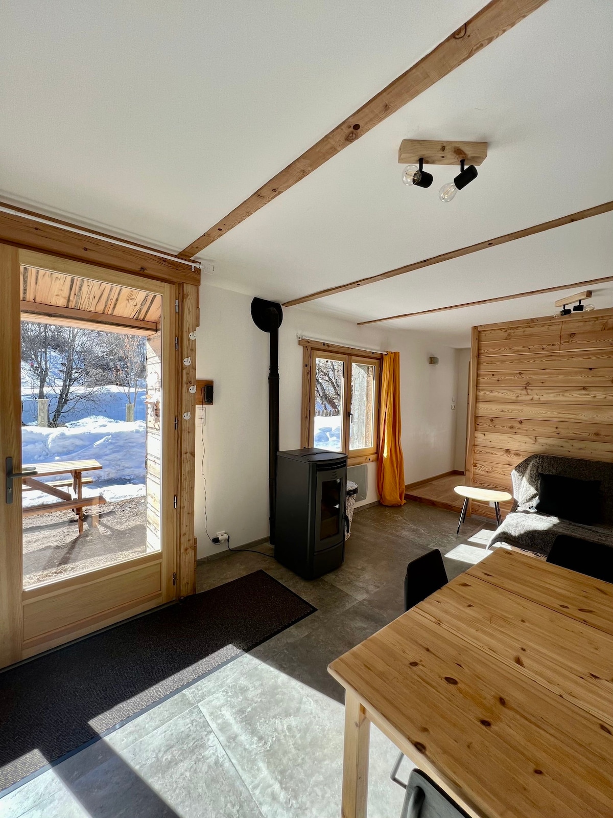 A cozy interior space featuring wooden accents and a large window allowing natural light. A modern wood stove provides warmth, while a wooden dining table and simple chairs invite meals together. The view outside shows a snowy landscape, enhancing the serene atmosphere.