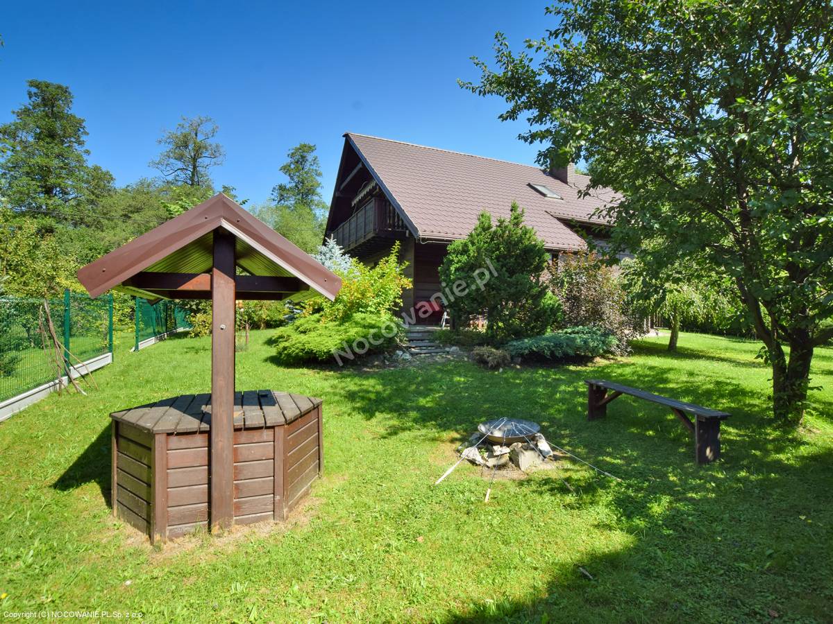 The exterior of a charming house is surrounded by lush greenery and features a cozy fire pit with a wooden bench. A traditional wooden well is positioned nearby, complemented by well-maintained shrubs and trees, creating a welcoming outdoor space.