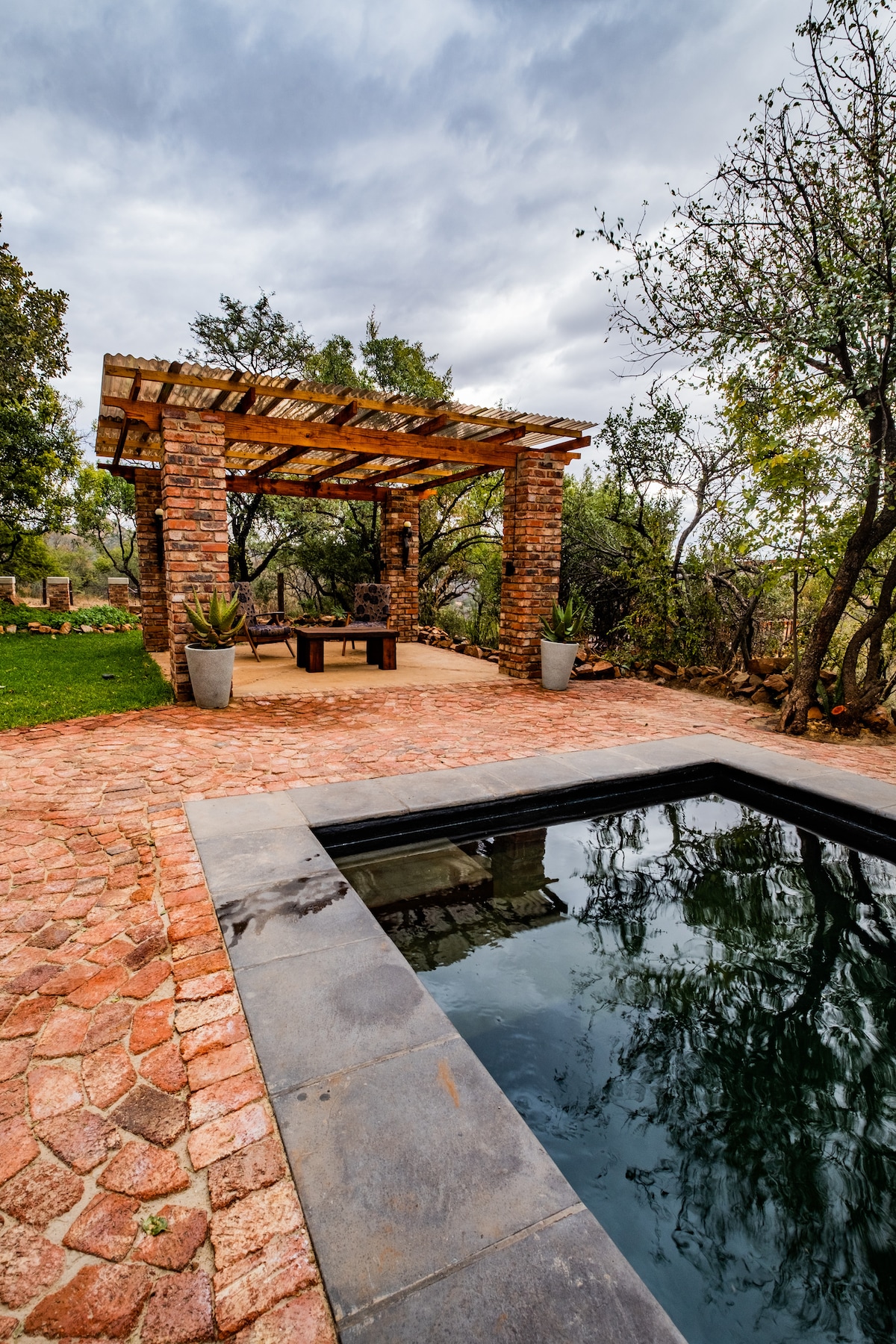 An outdoor area is showcased with a stone-paved patio, featuring a shaded pergola supported by rustic columns. A table is placed beneath the structure, surrounded by greenery. A small, still pool is visible nearby, with the surrounding landscape in a tranquil setting.