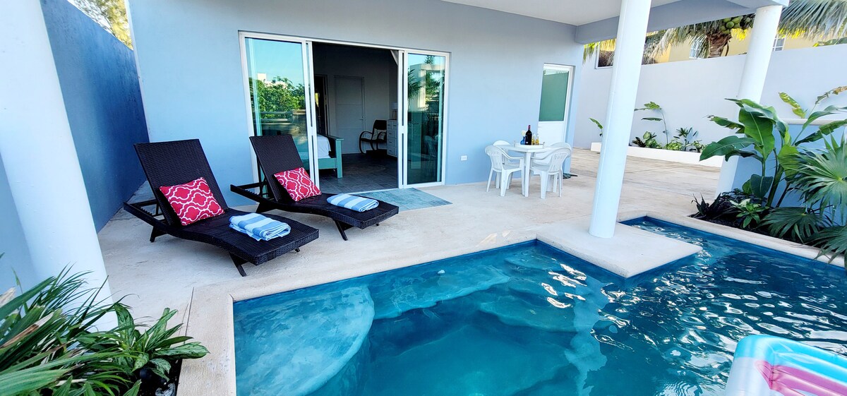 A private terrace features two lounge chairs with decorative pillows, positioned beside a filtered pool. A small dining table is set to the side, surrounded by tropical plants. Sliding glass doors provide access to the interior, enhancing the space's open-air feel.