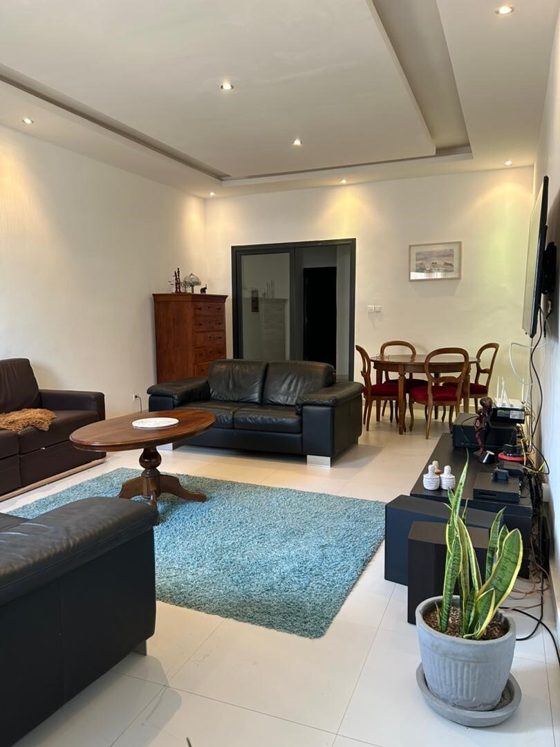 A well-lit living area features a cozy arrangement of leather sofas and a wooden coffee table on a soft rug. A dining table with four chairs is visible in the background, alongside a wooden cabinet and decorative items. Green plants add a touch of nature.
