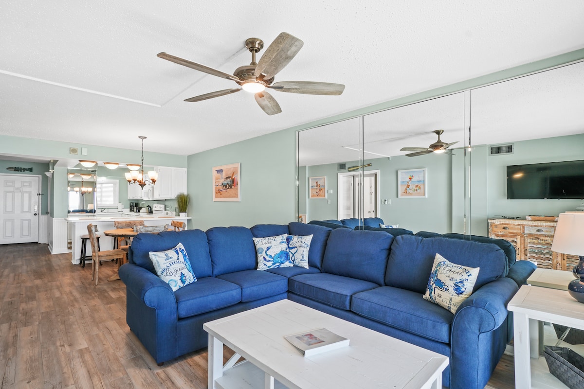 A spacious living area features a blue sectional sofa adorned with decorative pillows, complemented by a white coffee table. A ceiling fan is mounted above, and mirrored panels enhance the openness of the room. In the background, a dining area and kitchen are visible with light-colored furnishings.
