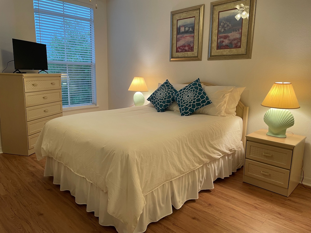 A comfortable sleeping area is featured with a spacious bed adorned in crisp white linens. Two bedside tables with lamps provide soft lighting, alongside decorative cushions. A small dresser and a television are positioned nearby, while natural light filters through the window with sheer curtains.
