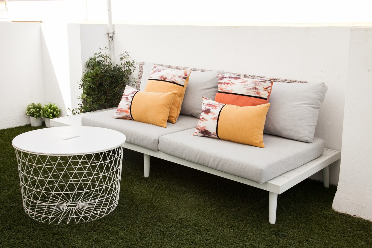 A comfortable outdoor seating area features a light-colored sofa adorned with colorful cushions in shades of yellow and orange. A round white table with a geometric design complements the setup, set on a grassy surface surrounded by potted greenery.