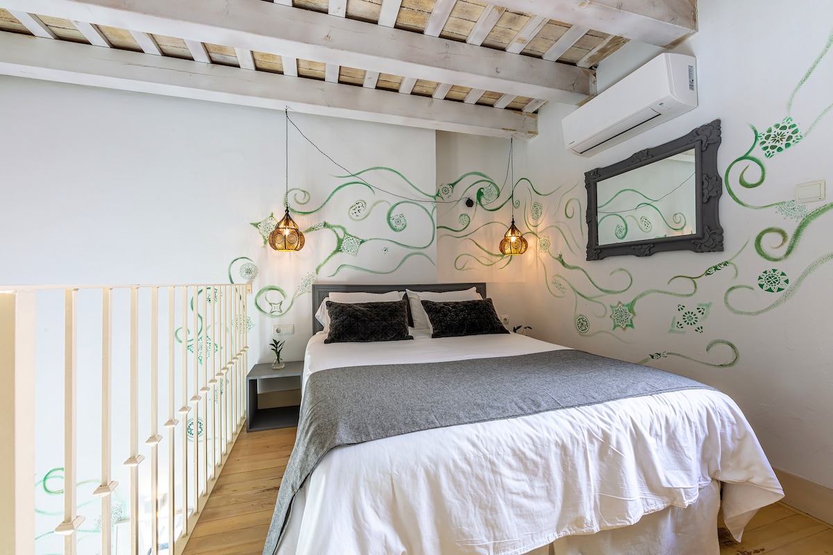A cozy sleeping area is featured with a spacious bed, adorned with a light blanket. Unique wall art in green swirls decorates the white walls. Two pendant lights hang above the bed, while a mirror is positioned on the nearby wall. Wood beams add character to the ceiling.