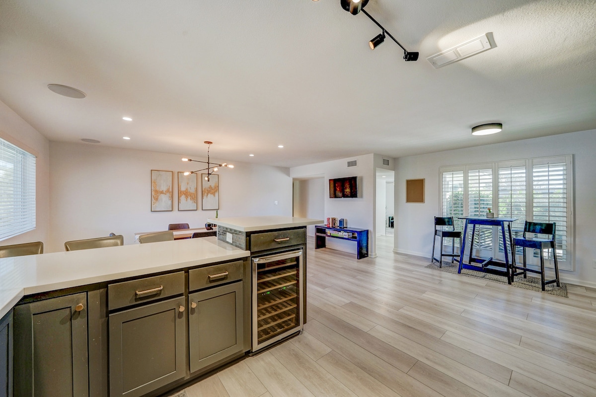 A spacious living area features a modern kitchen island with a built-in wine cooler. Light-colored wood flooring extends throughout the room. Large windows allow plenty of natural light, while minimalist furniture and art pieces create a clean and contemporary aesthetic.