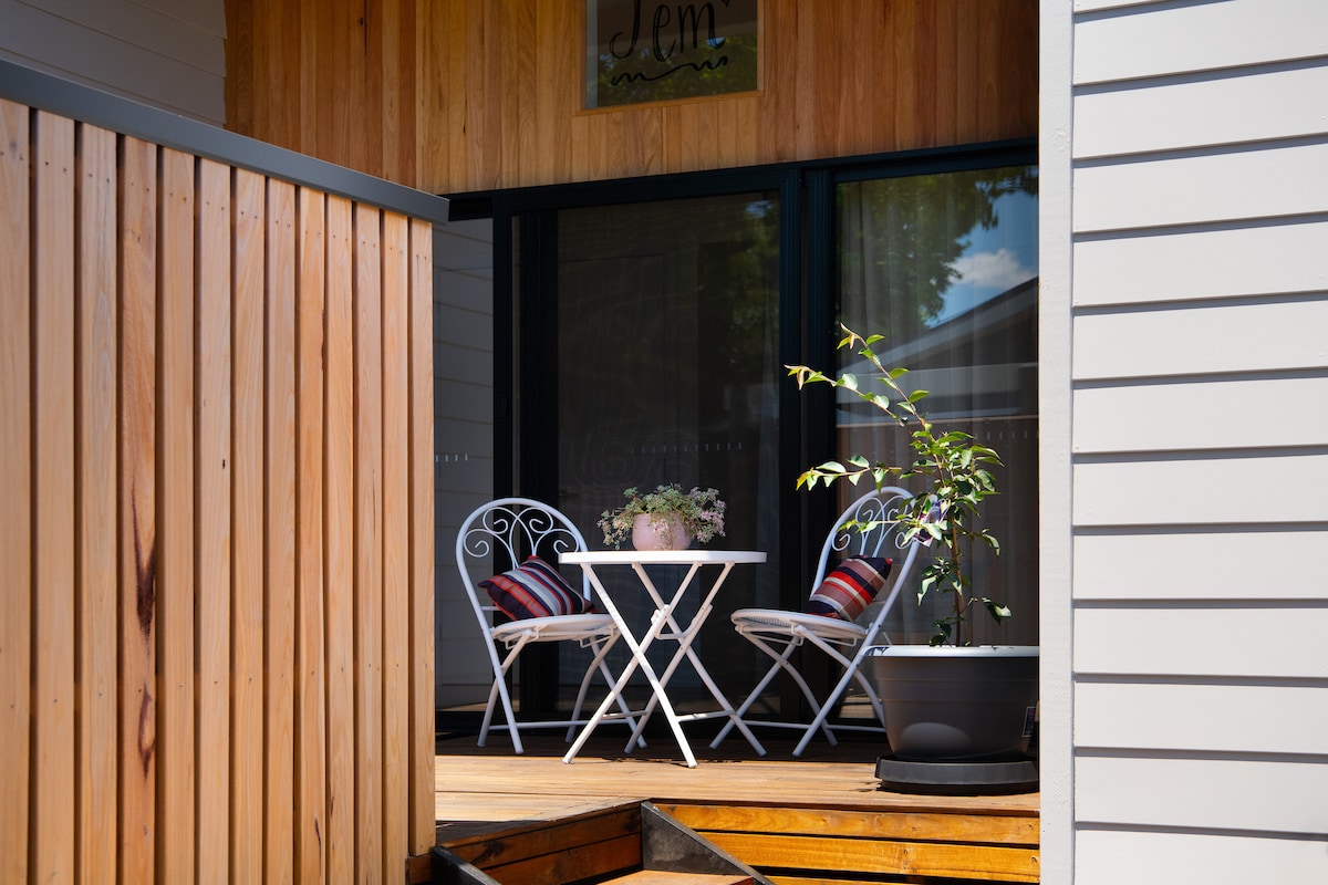 A cozy outdoor seating area is highlighted by a small round table and two white metal chairs adorned with colorful cushions. A potted plant adds a touch of greenery, while the wooden deck and textured wooden paneling create a warm entrance.