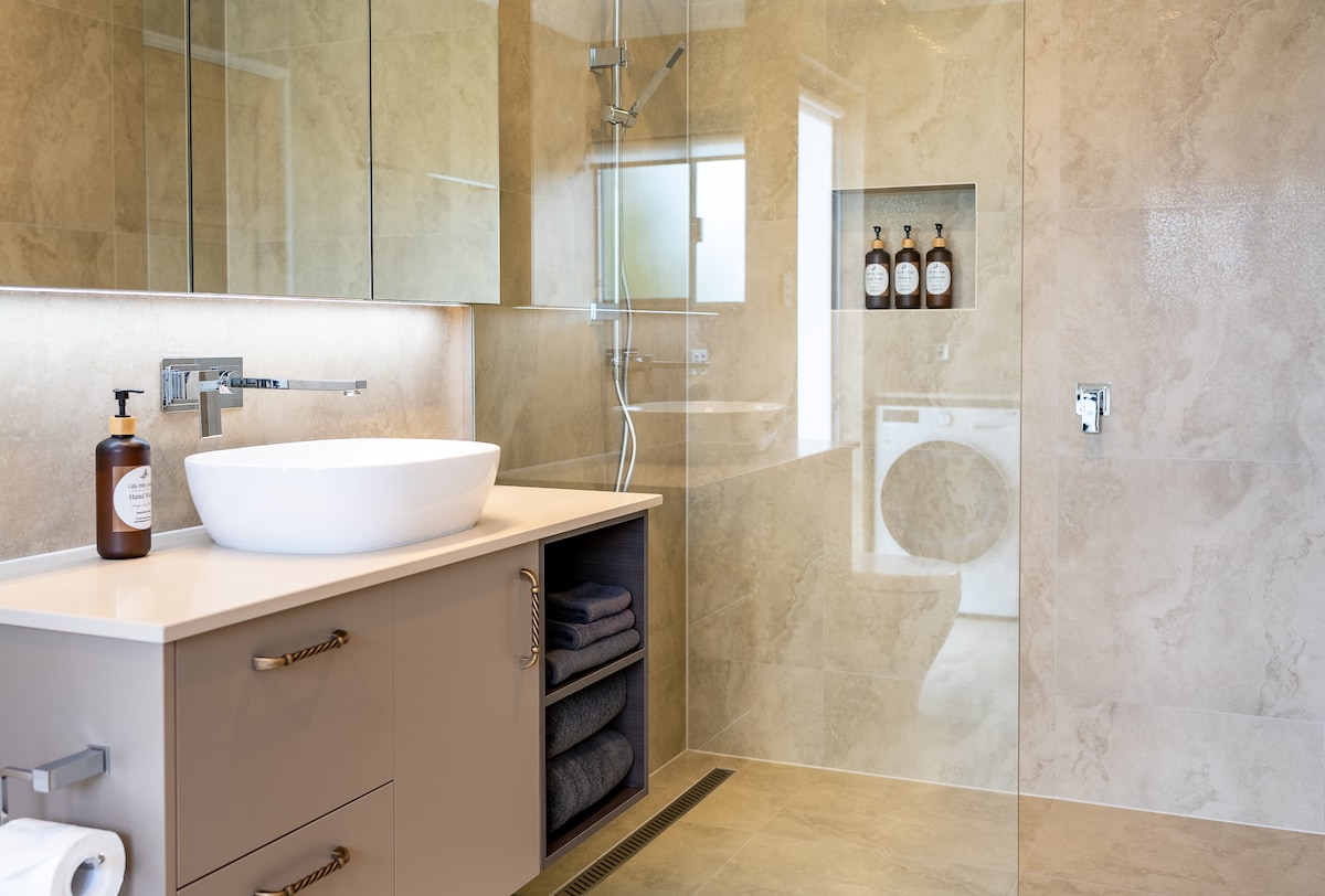 A modern bathroom features a round vessel sink atop a sleek vanity with drawers. neutral stone tiles cover the walls and floor. A spacious shower is enclosed with glass panels, and laundry facilities are visible in the background.