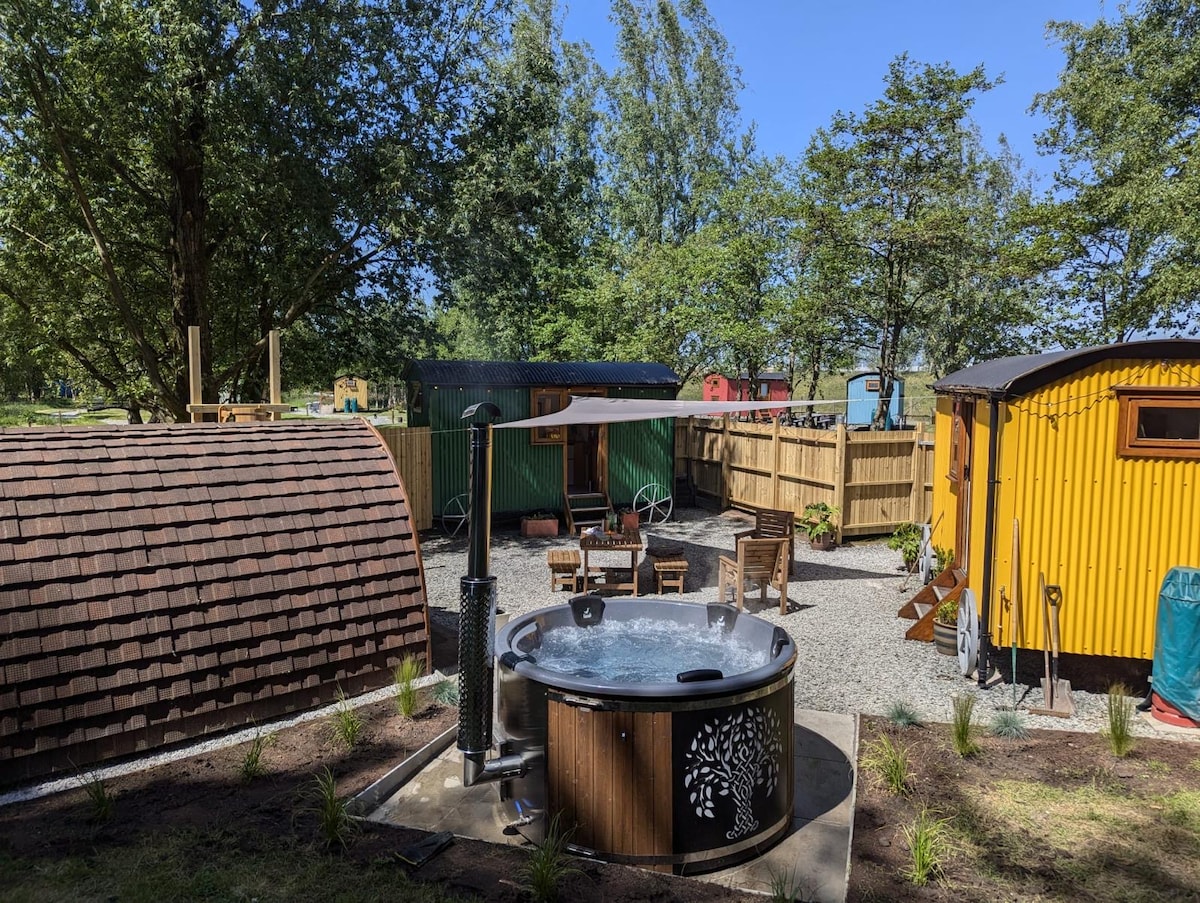 A wood burning hot tub is positioned at the forefront, surrounded by lush greenery and a gravel pathway. Nearby, colorful shepherd huts are visible, adding a cozy charm to the outdoor space. Sunlight filters through the trees, enhancing the serene atmosphere.