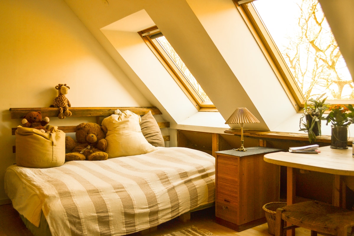 A light-filled sleeping area features a spacious bed dressed in a neutral-toned blanket. Soft stuffed animals are arranged at the head of the bed. Angled windows provide natural light, while a small lamp and a flower arrangement sit on a side table nearby.