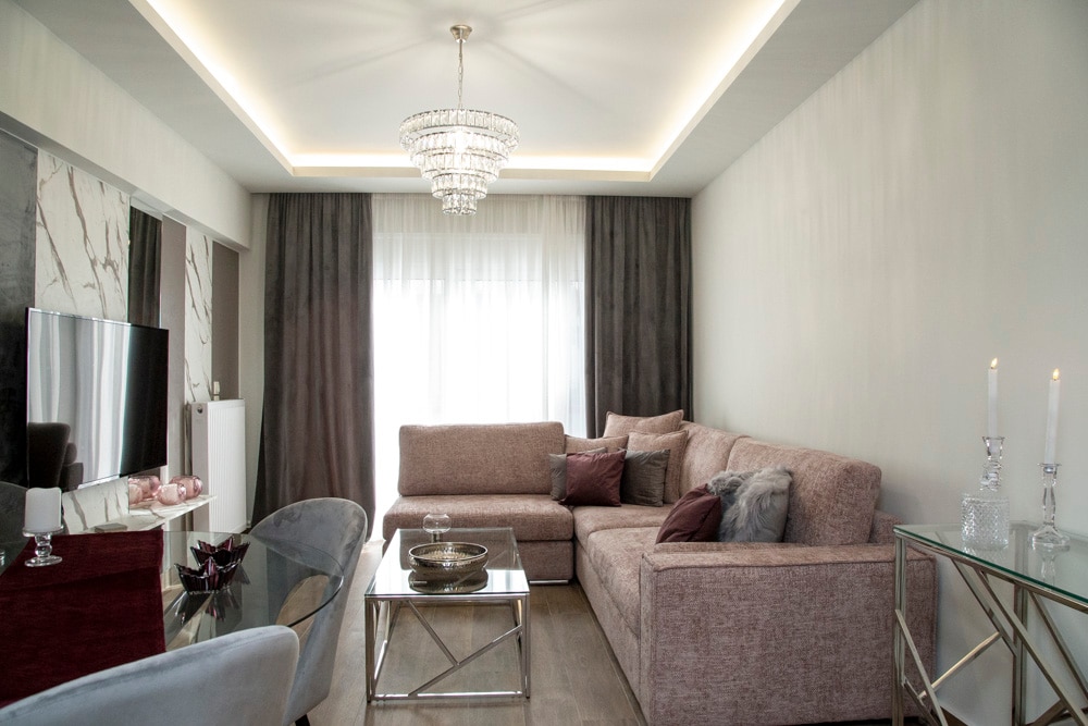 A contemporary living area is highlighted by a plush sectional sofa adorned with decorative pillows. Natural light filters through sheer curtains, illuminating a modern coffee table with elegant decor. A ceiling chandelier adds a touch of sophistication, complemented by a stylish TV unit against a marble accent wall.