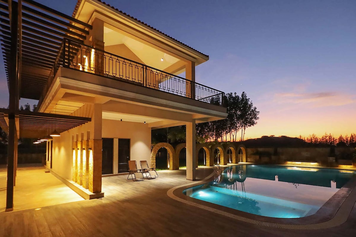 The private outdoor pool is framed by a spacious deck, showcasing comfortable lounge chairs. The structure, adorned with elegant arches, features soft lighting that enhances the tranquil ambiance during sunset. Lush greenery is visible in the background, complementing the serene environment.