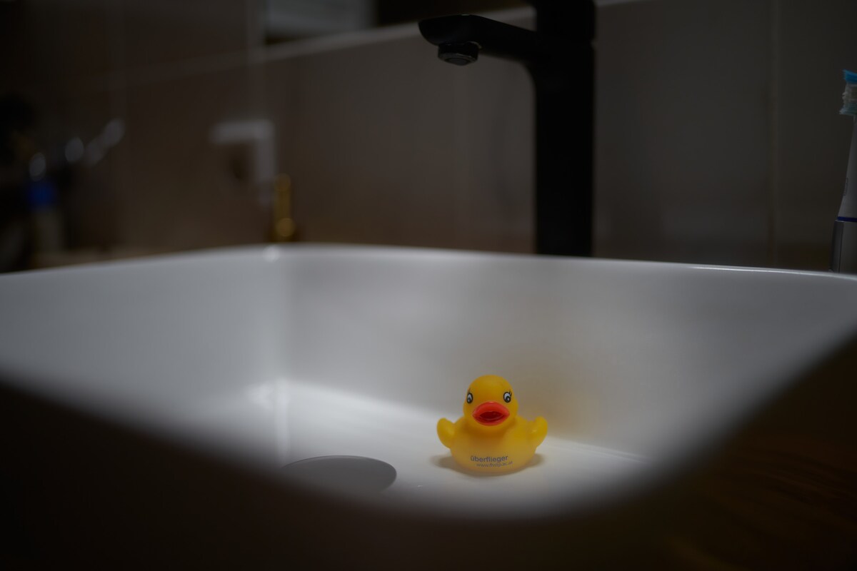 A white sink is showcased, with a small yellow rubber duck floating in the center. The sink's sleek design is highlighted by the surrounding soft light, creating a clean and minimalistic atmosphere.