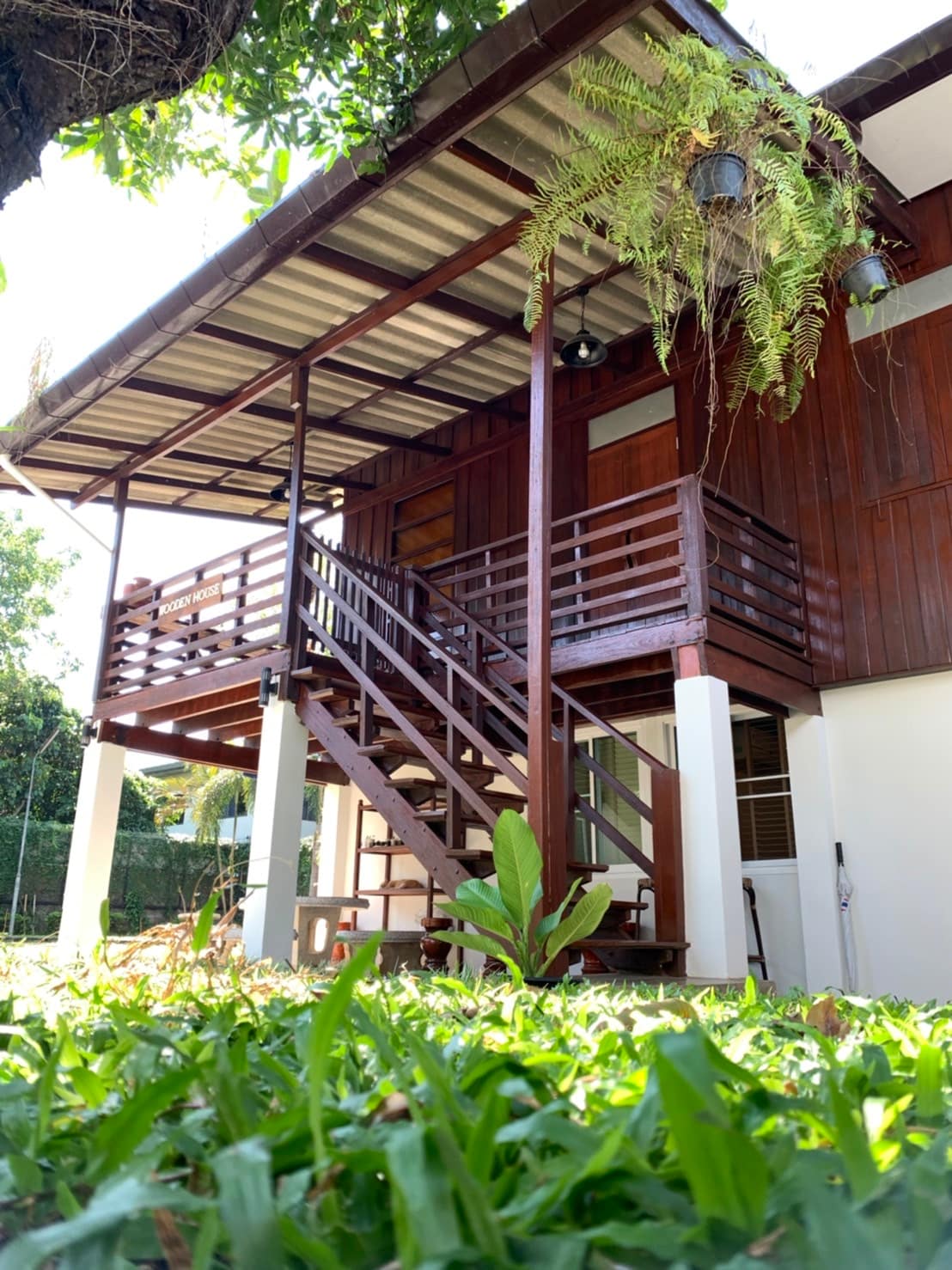 A two-story wooden house is depicted with a staircase leading to a spacious balcony. The structure features a modern design with natural wood tones, set against a backdrop of lush green grass and trees. Hanging plants add a touch of nature, enhancing the inviting exterior.