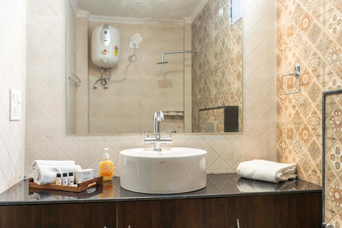 A modern bathroom features a sleek round sink atop a dark countertop. A large mirror reflects the intricate patterned tiles on the wall. Toiletries and towels are neatly arranged on the counter, enhancing the tidy and functional atmosphere.