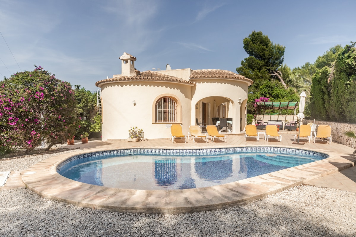 A villa with a rounded design features a private pool surrounded by a sun terrace and lounge chairs. Lush greenery and flowering plants add to the tranquil setting, while the exterior showcases a smooth stucco finish under a clear sky.