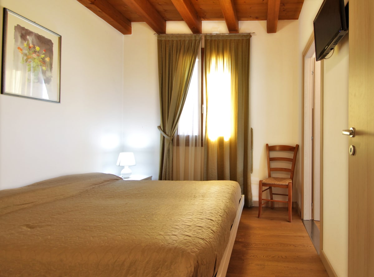 A bright sleeping area features a spacious bed with neutral bedding. A small lamp illuminates a bedside table beside the bed. Natural light enters through a window, dressed with green curtains, and a wooden chair adds a touch of warmth to the room.