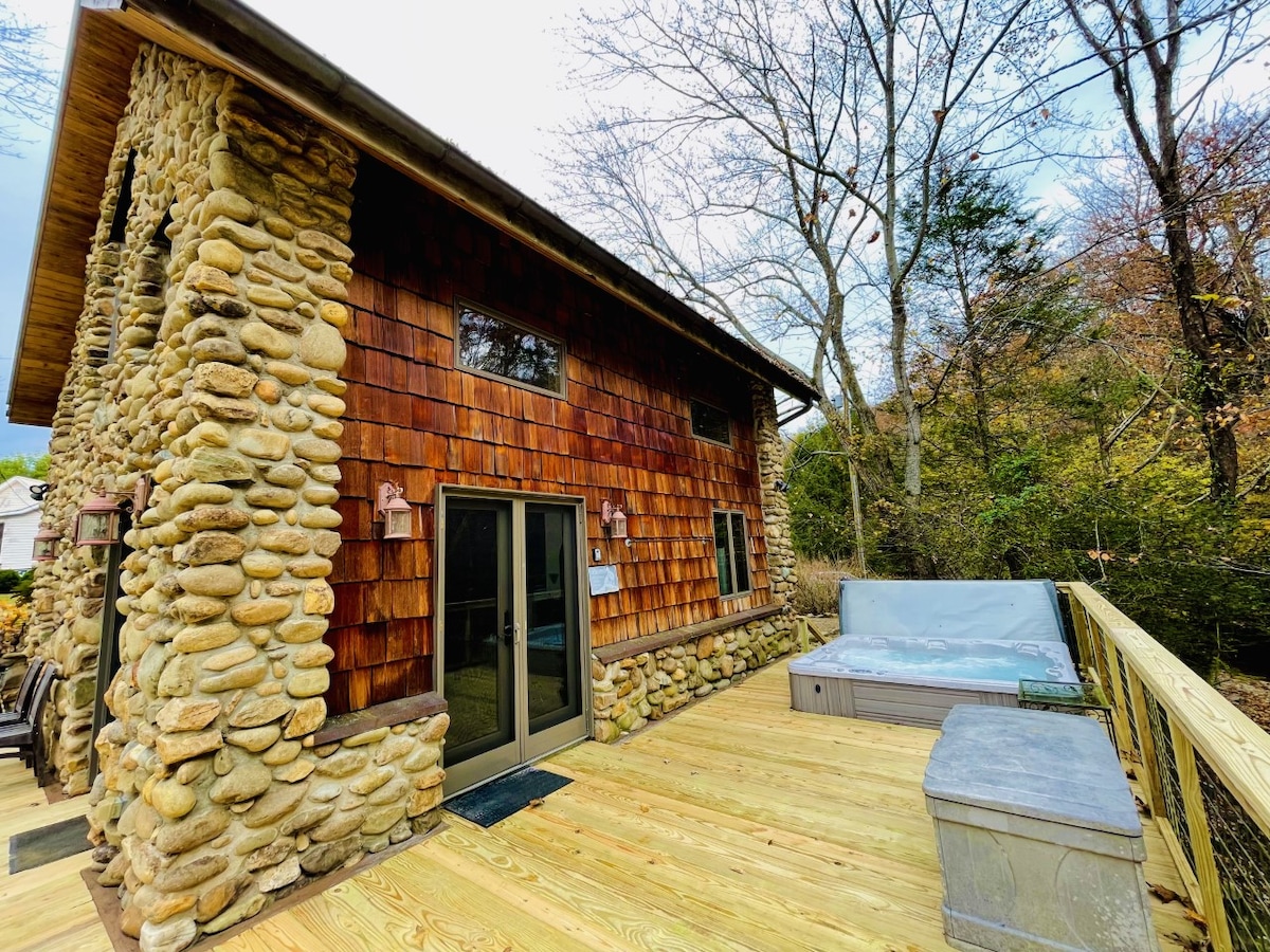 The exterior of the Stone Cottage showcases a blend of natural stone and wooden cedar shakes. A spacious deck extends from the cottage, featuring a jacuzzi tub and steps leading to the surrounding nature. Large glass doors provide access to the indoors.