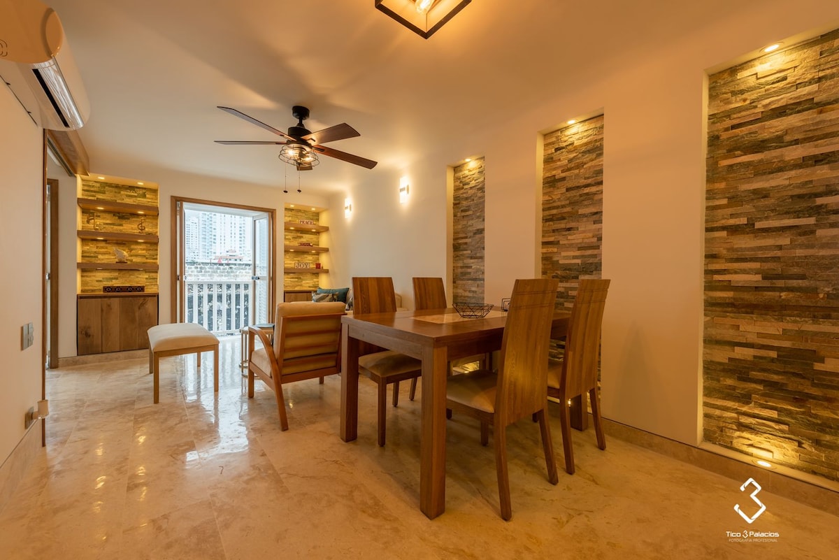 A warm dining area features a wooden table surrounded by six chairs, complemented by a nearby bench. Natural light filters through the balcony door, accentuating the textured stone walls and soft ambient lighting. A ceiling fan adds a functional touch to the inviting space.