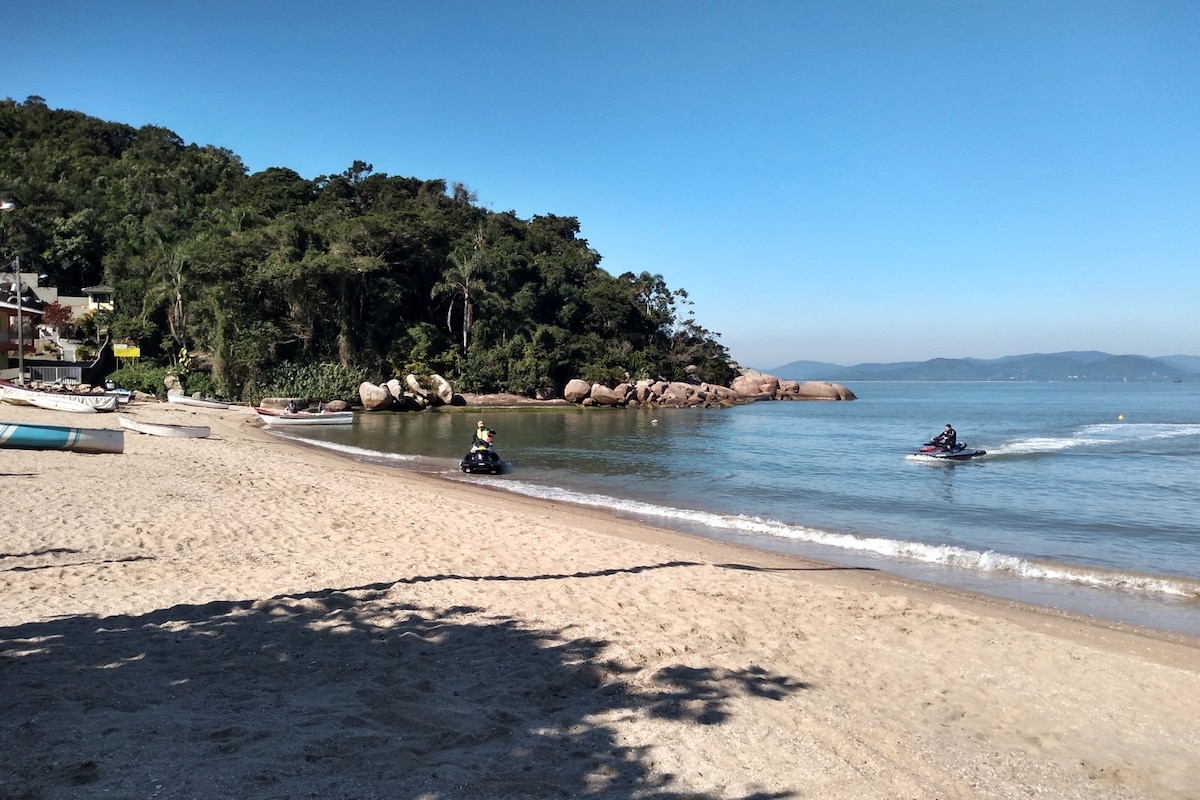 A sandy beach is seen stretching along the water's edge, with gentle waves lapping at the shore. Lush greenery lines the background, complementing smooth rocks dotting the landscape. Two jet skis are visible on the water, hinting at recreational activities available in the area.