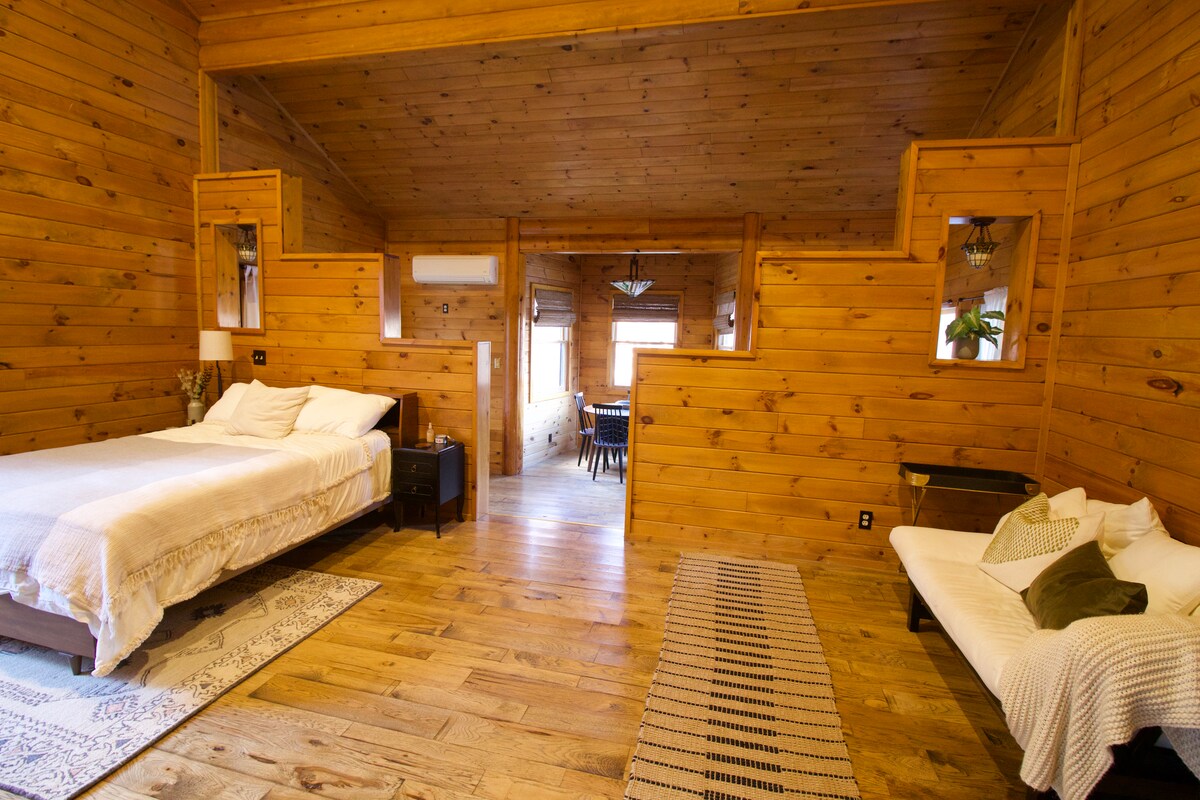 The open studio space features warm wooden paneling and high ceilings. A spacious bed with neutral linens is situated beside a nightstand. A daybed is positioned to the right, with a light rug underneath, leading towards a cozy dining area visible through the doorway.