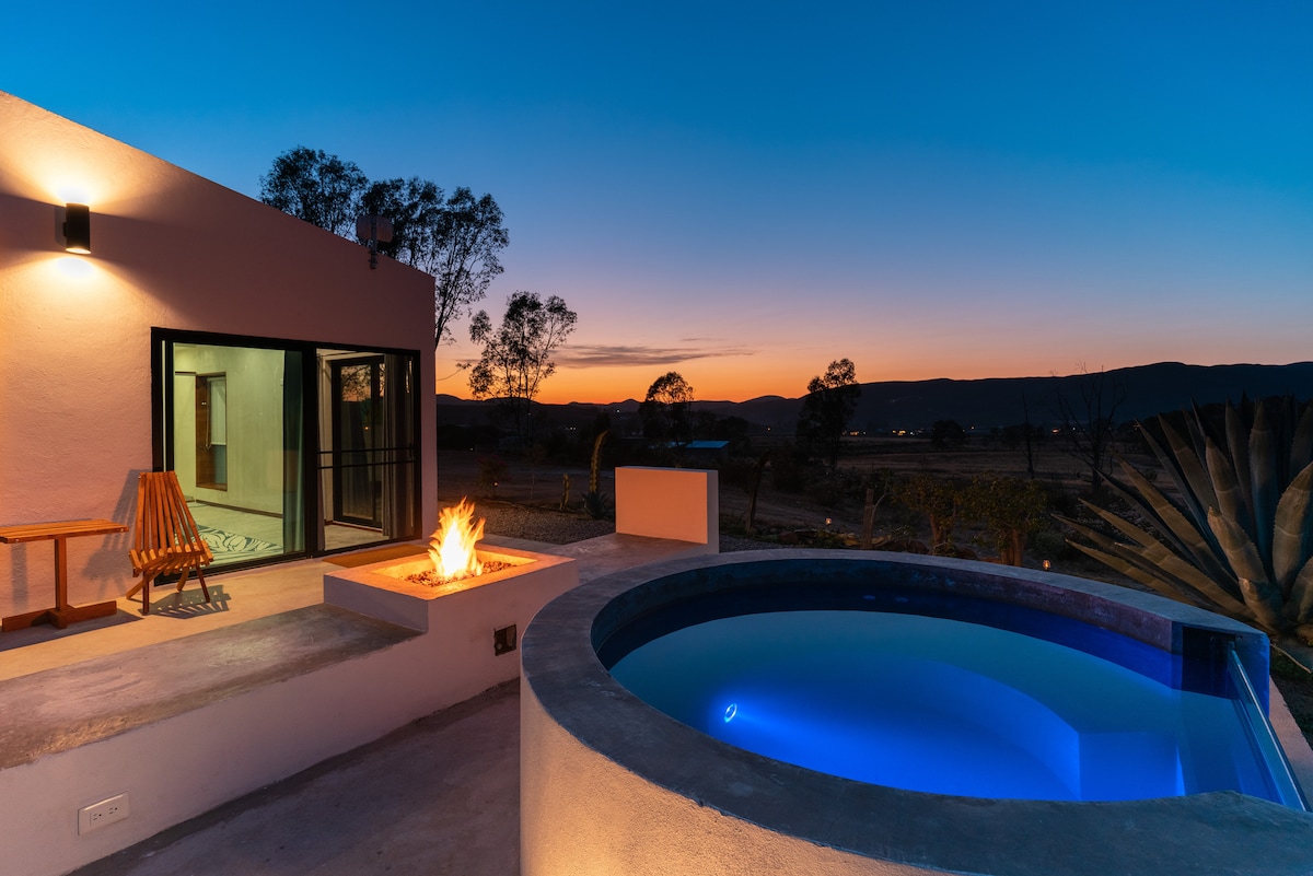An outdoor area features a private jacuzzi with illuminated water, complemented by a fire pit nearby. The setting is framed by gentle hills under a twilight sky, creating a serene atmosphere for relaxation and enjoyment.