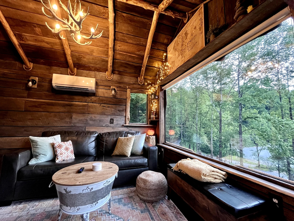 The living room features a large, dark sofa accented by decorative pillows and a coffee table with a cozy texture. A floor-to-ceiling window offers views of the wooded surroundings, while string lights and an antler chandelier provide warm illumination.