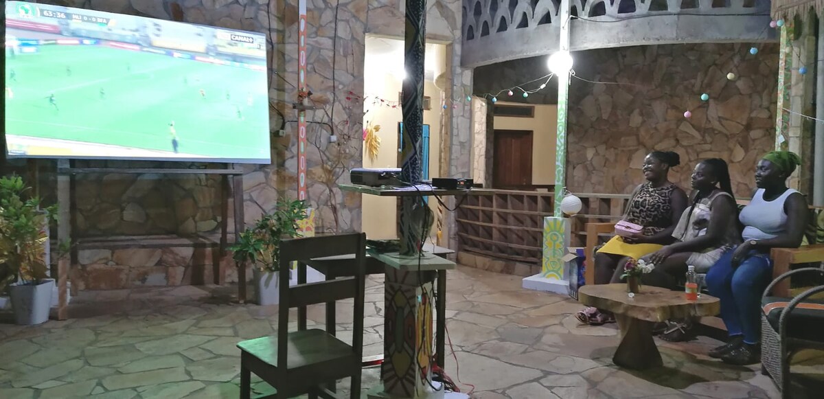 An outdoor seating area features a large screen displaying a sports event. Comfortable seating arrangements, including a small wooden table and chairs, are positioned around the space. Stone walls and decorative lighting add to the inviting environment, creating an ideal spot for gathering.